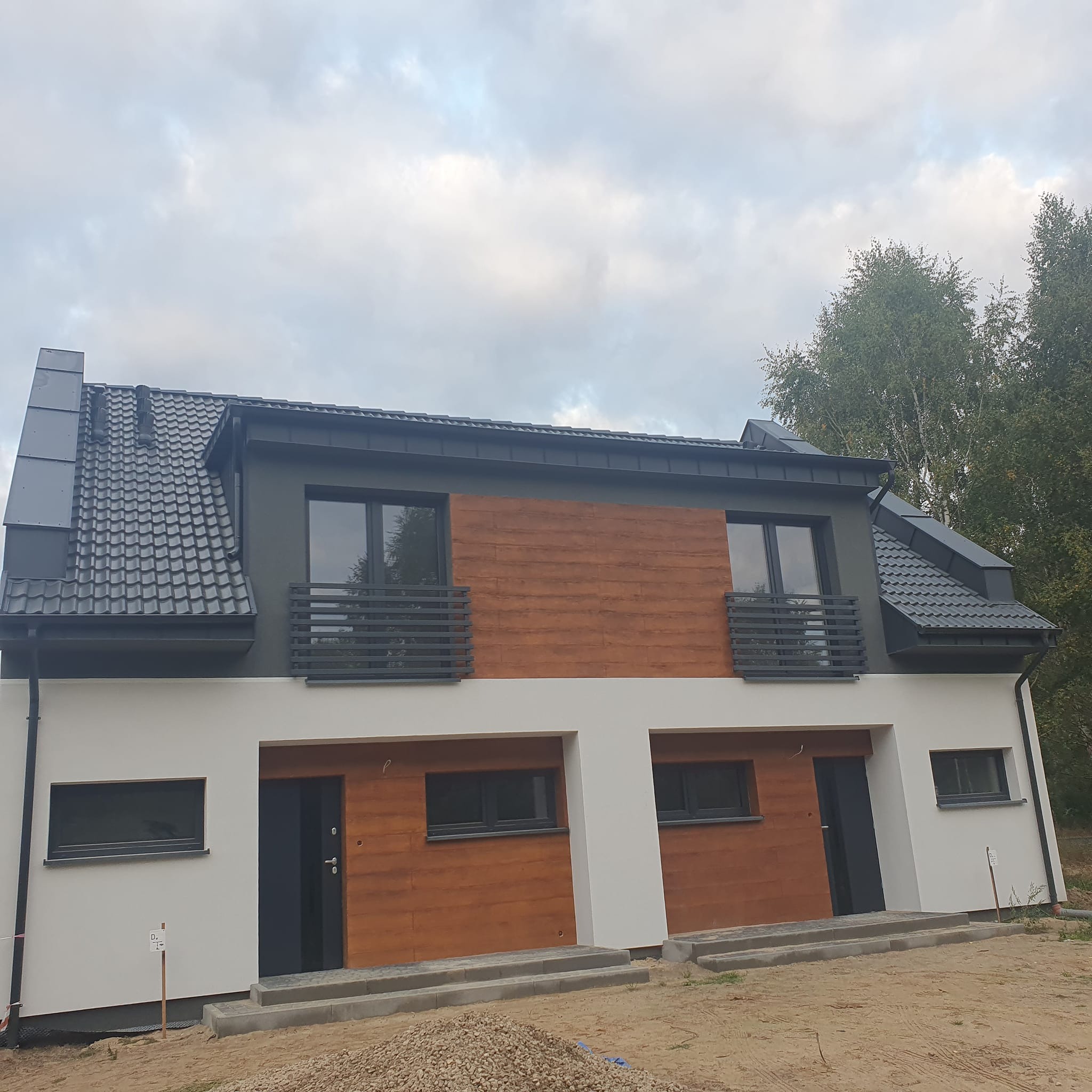 Nowoczesny bliźniak z grafitową dachówką i drewnianą elewacją. Widoczne rynny, okna i drzwi w kolorze antracytowym. Tło: pochmurne niebo i drzewa.