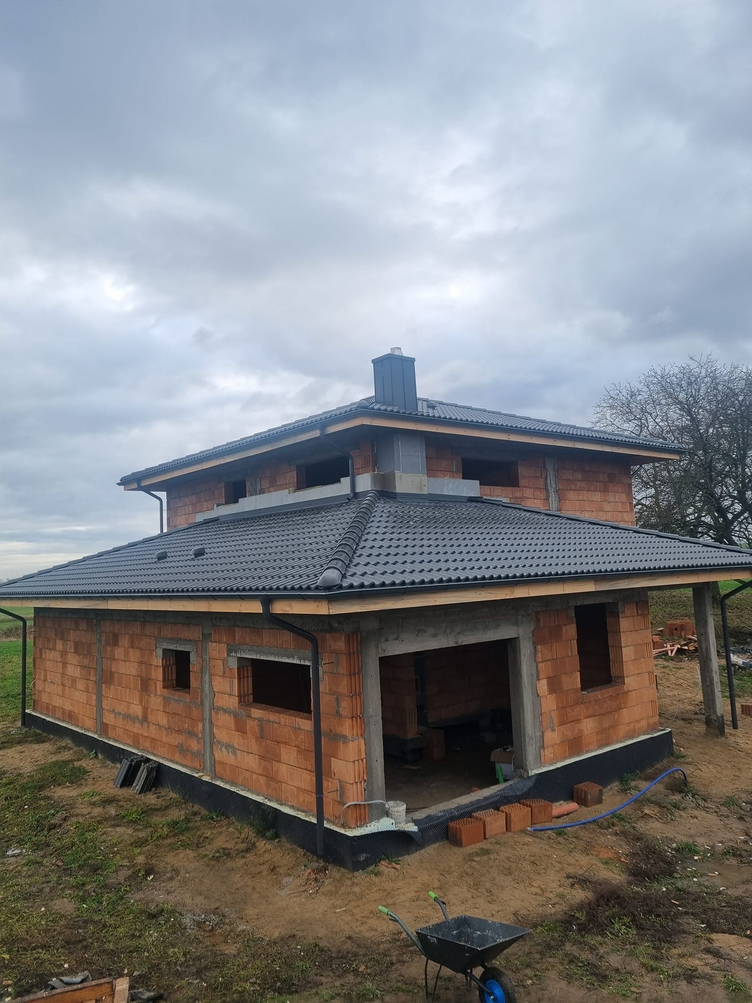 Dwukondygnacyjny dom w trakcie budowy z częściowo pokrytym dachem z czarnej dachówki, widoczne pustaki i rynny, na pierwszym planie taczka.