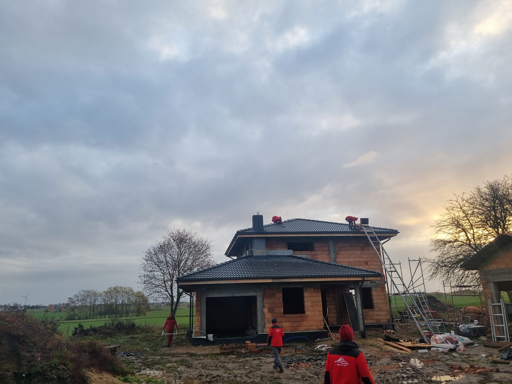Dom w trakcie budowy z czarnym dachem z dachówki, pracownicy w czerwonych ubraniach na dachu i wokół budynku. Pochmurne niebo w tle, krajobraz wiejski.