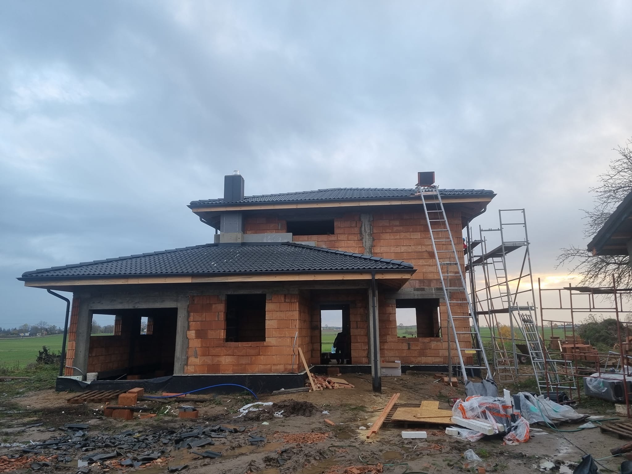 Dom w trakcie budowy z częściowo położonym, ciemnym pokryciem. Widoczne rusztowania i drabina. Otoczenie placu budowy, pochmurne niebo.