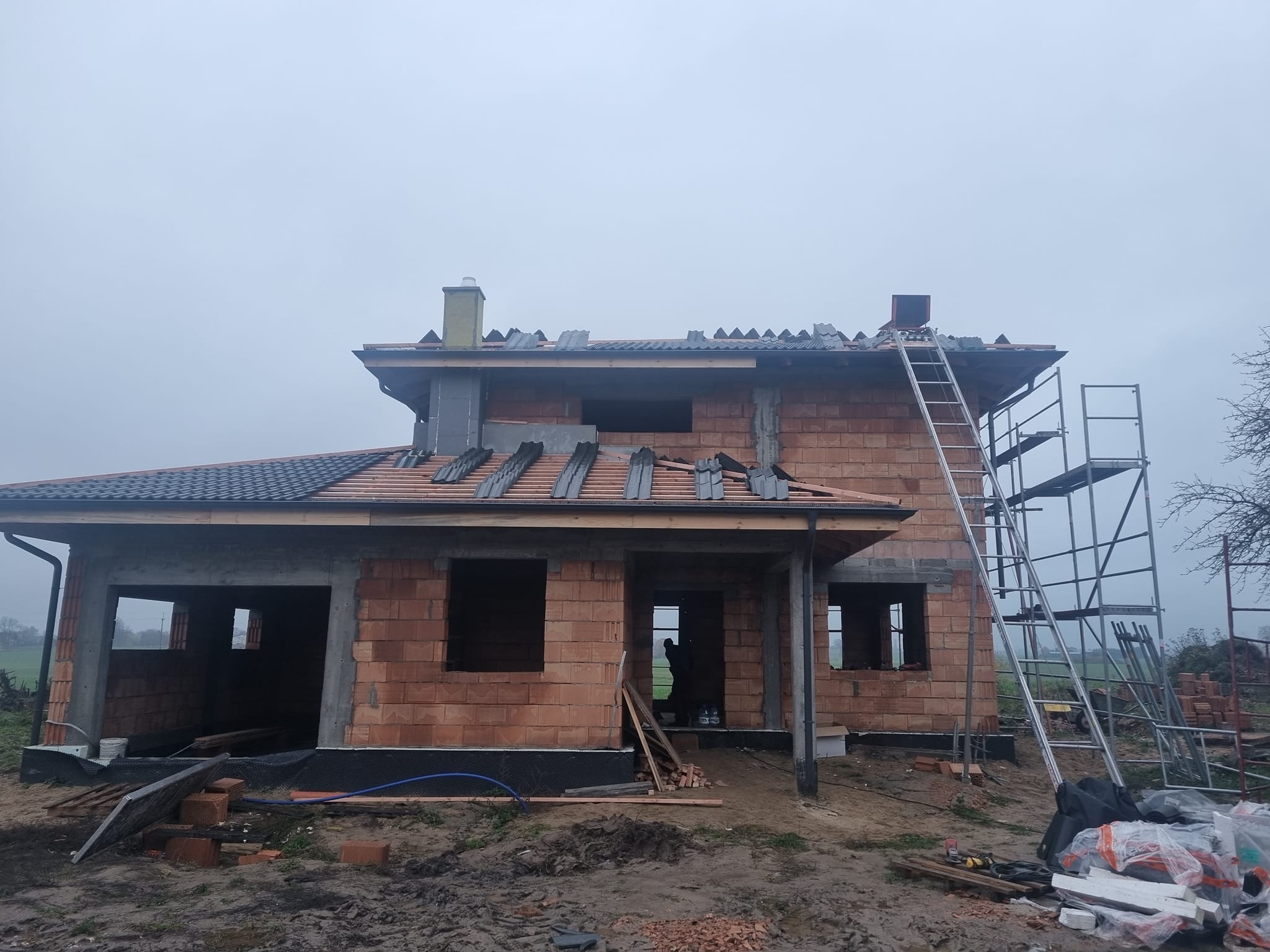 Budowa domu jednorodzinnego w trakcie prac dekarskich: częściowo położone dachówki, ceglana konstrukcja, rusztowanie. Widok na budynek w pochmurny dzień.