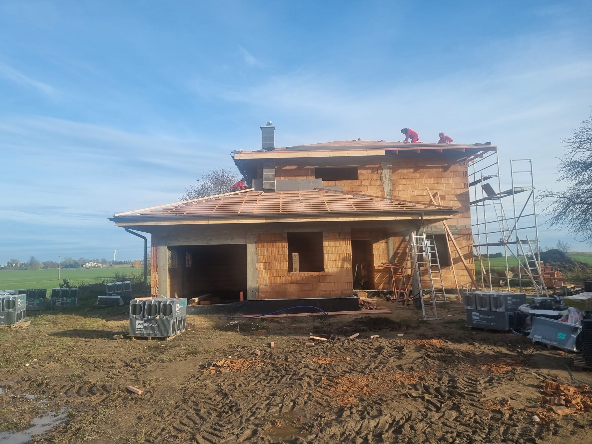 Budowa domu jednorodzinnego w trakcie krycia dachu dachówką ceramiczną, widoczni dekarze pracujący na drewnianej konstrukcji, palety z dachówkami na ziemi.