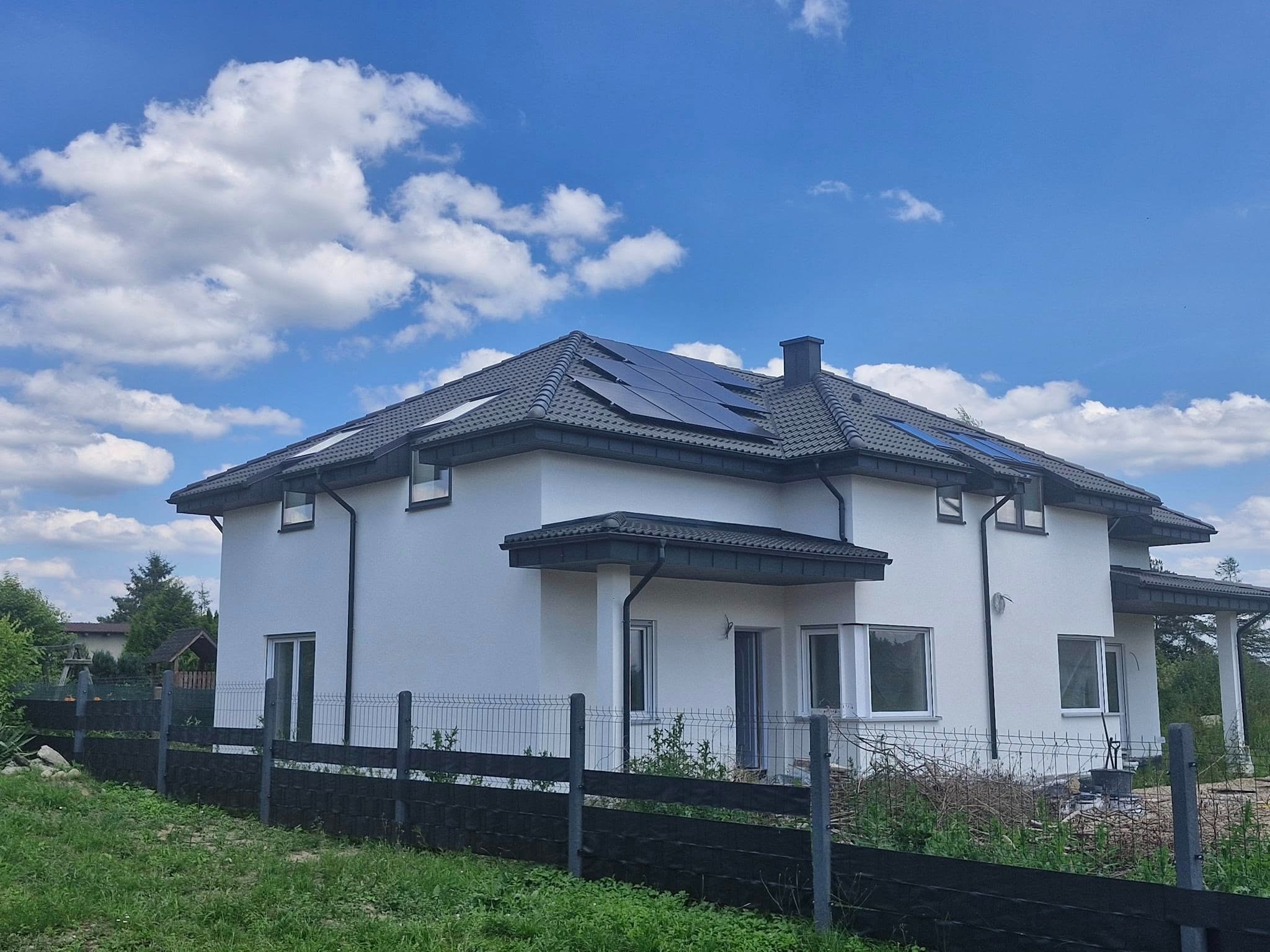 Biały dom jednorodzinny z szarym dachem i panelami słonecznymi, widziany z poziomu gruntu, ogrodzony czarnym płotem na tle błękitnego nieba z chmurami.