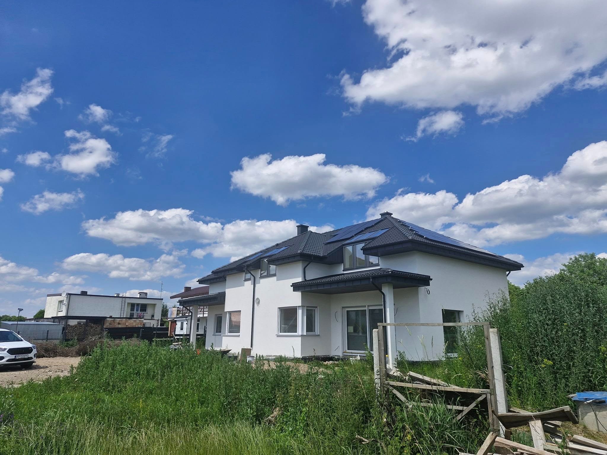 Nowoczesny dom z ciemną dachówką i panelami słonecznymi na dachu, na tle błękitnego nieba z chmurami. Widoczne elementy ogrodu i sąsiednie budynki.