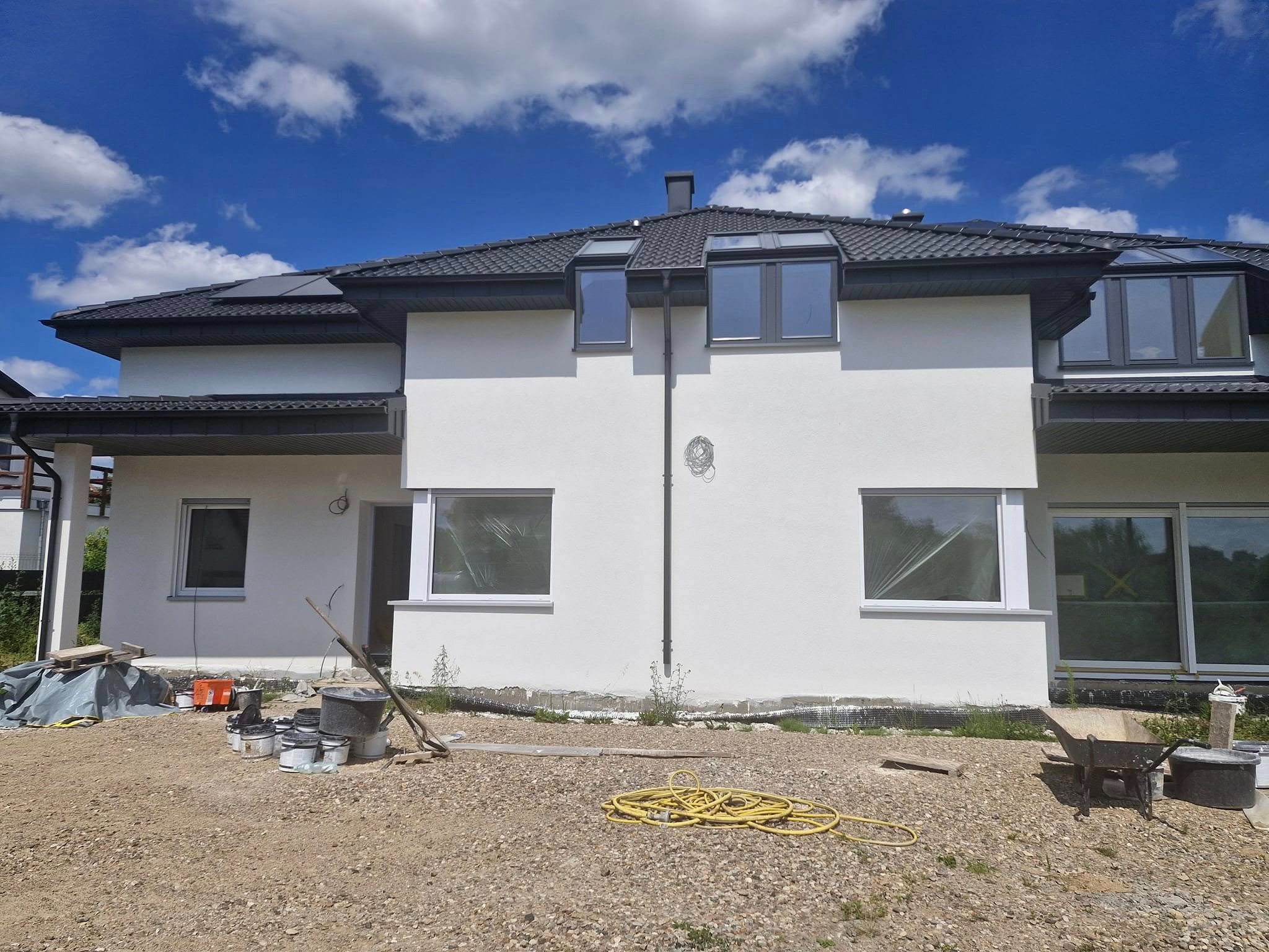 Dwupiętrowy dom z ciemną dachówką i jasną elewacją w trakcie prac wykończeniowych. Na pierwszym planie widoczne narzędzia i materiały budowlane.