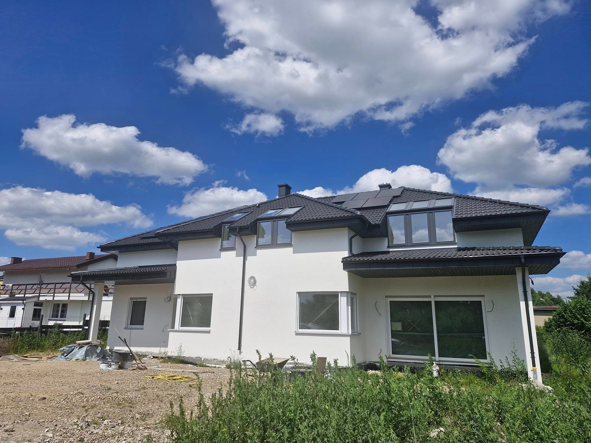 Nowoczesny dom jednorodzinny z ciemną dachówką i panelami słonecznymi na dachu, na tle błękitnego nieba z białymi chmurami. Widoczne narzędzia budowlane.