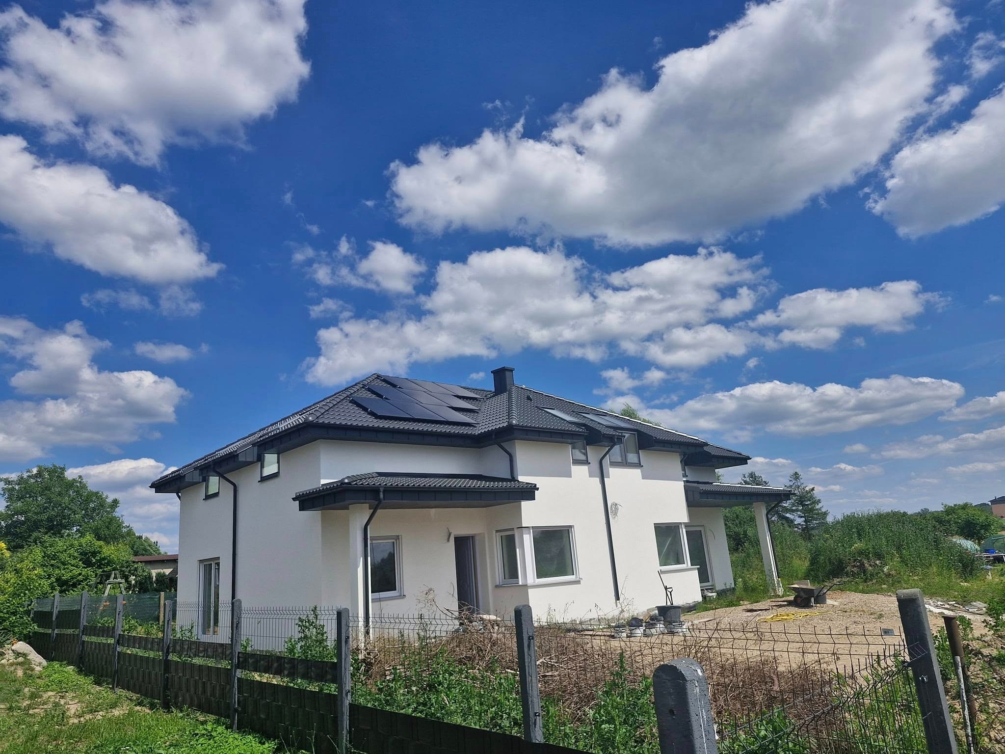 Nowoczesny, dwukondygnacyjny dom z ciemną dachówką i panelami solarnymi na dachu, widziany zza ogrodzenia, na tle błękitnego nieba z obłokami.