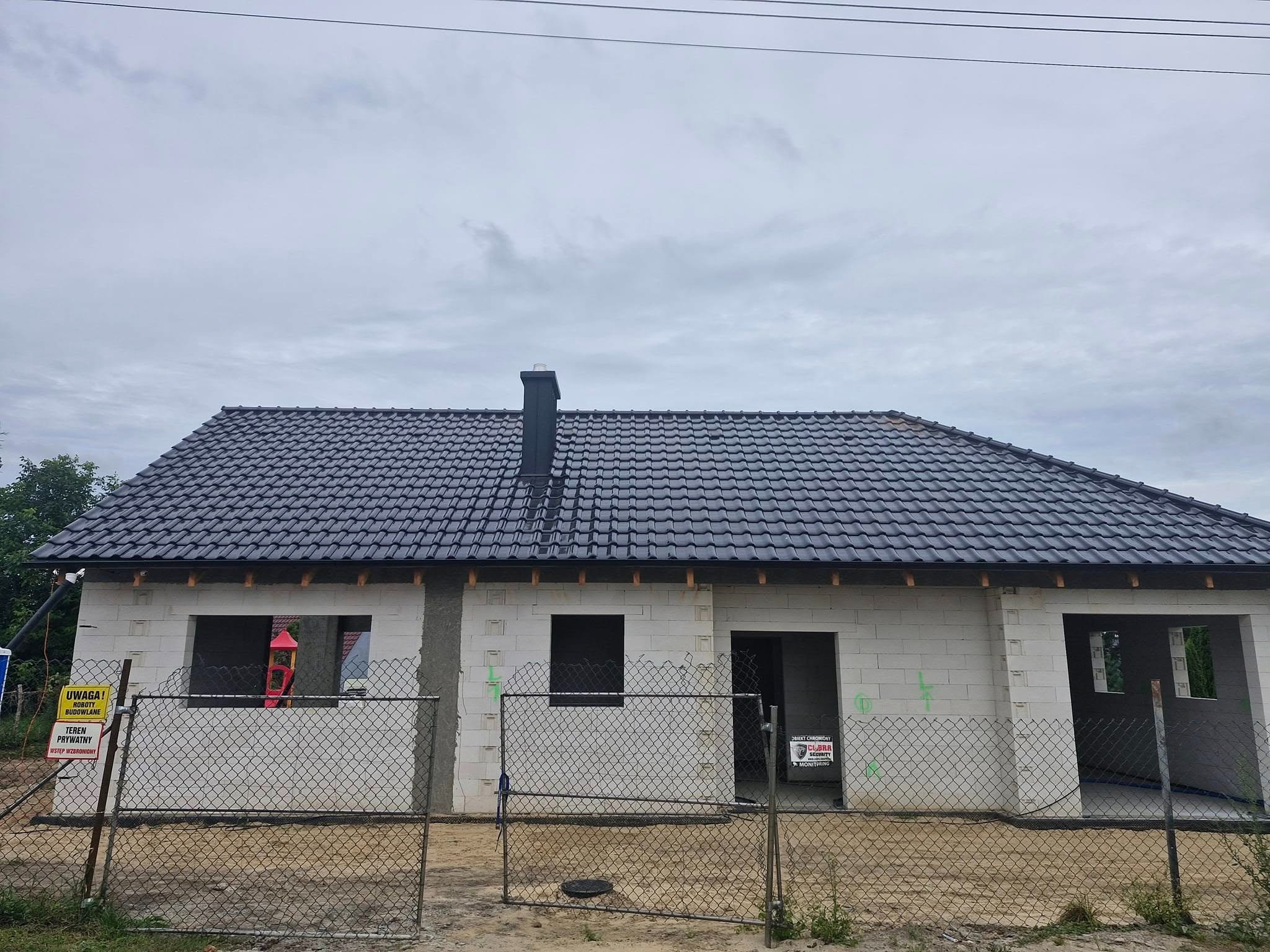 Dom w trakcie budowy z ciemnoszarą dachówką ceramiczną i czarnym kominem, ogrodzony siatką. Widoczne znaki ostrzegawcze.