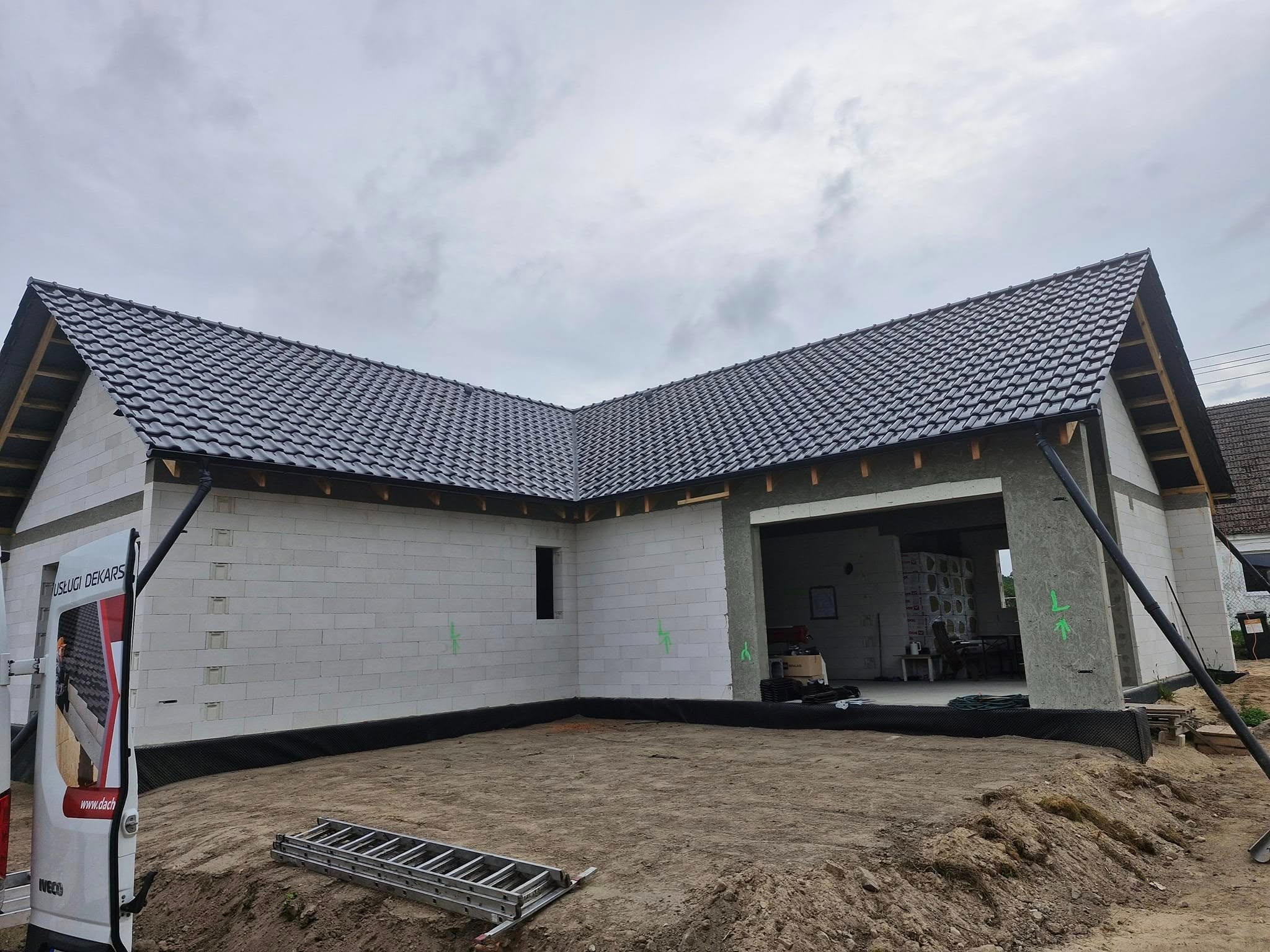 Dom w trakcie budowy z ciemną dachówką ceramiczną, widoczny fragment elewacji z białego betonu komórkowego, drabina i samochód dostawczy z logo firmy dekarskiej.