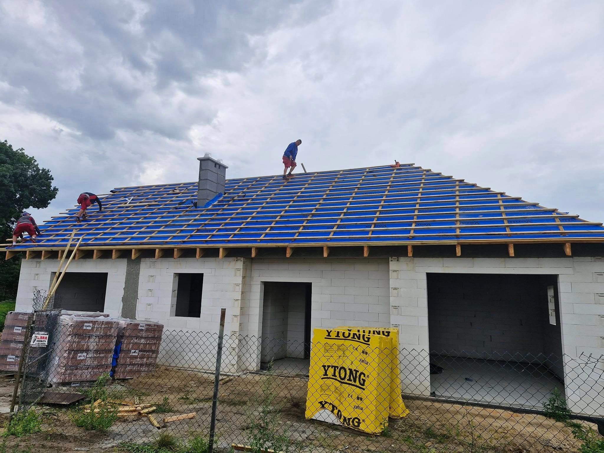 Ekipa montuje konstrukcję dachu na nowym domu z bloczków Ytong. Widoczna niebieska membrana i drewniane łaty. Na pierwszym planie palety z dachówką.