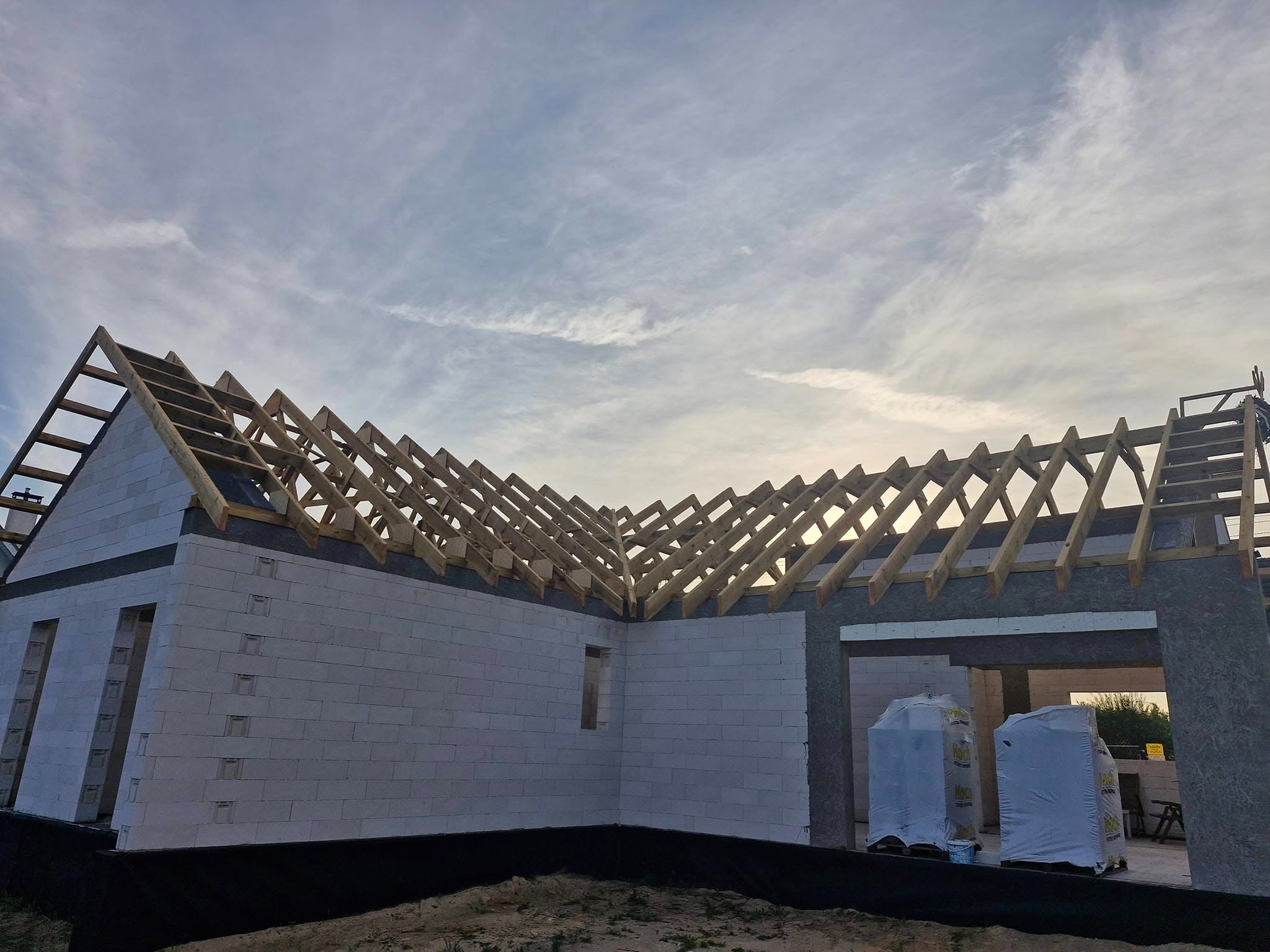 Wznoszenie konstrukcji dachu z drewnianą więźbą na murowanym budynku w stanie surowym otwartym. Widoczne pakiety materiałów budowlanych. Pochmurne niebo w tle.