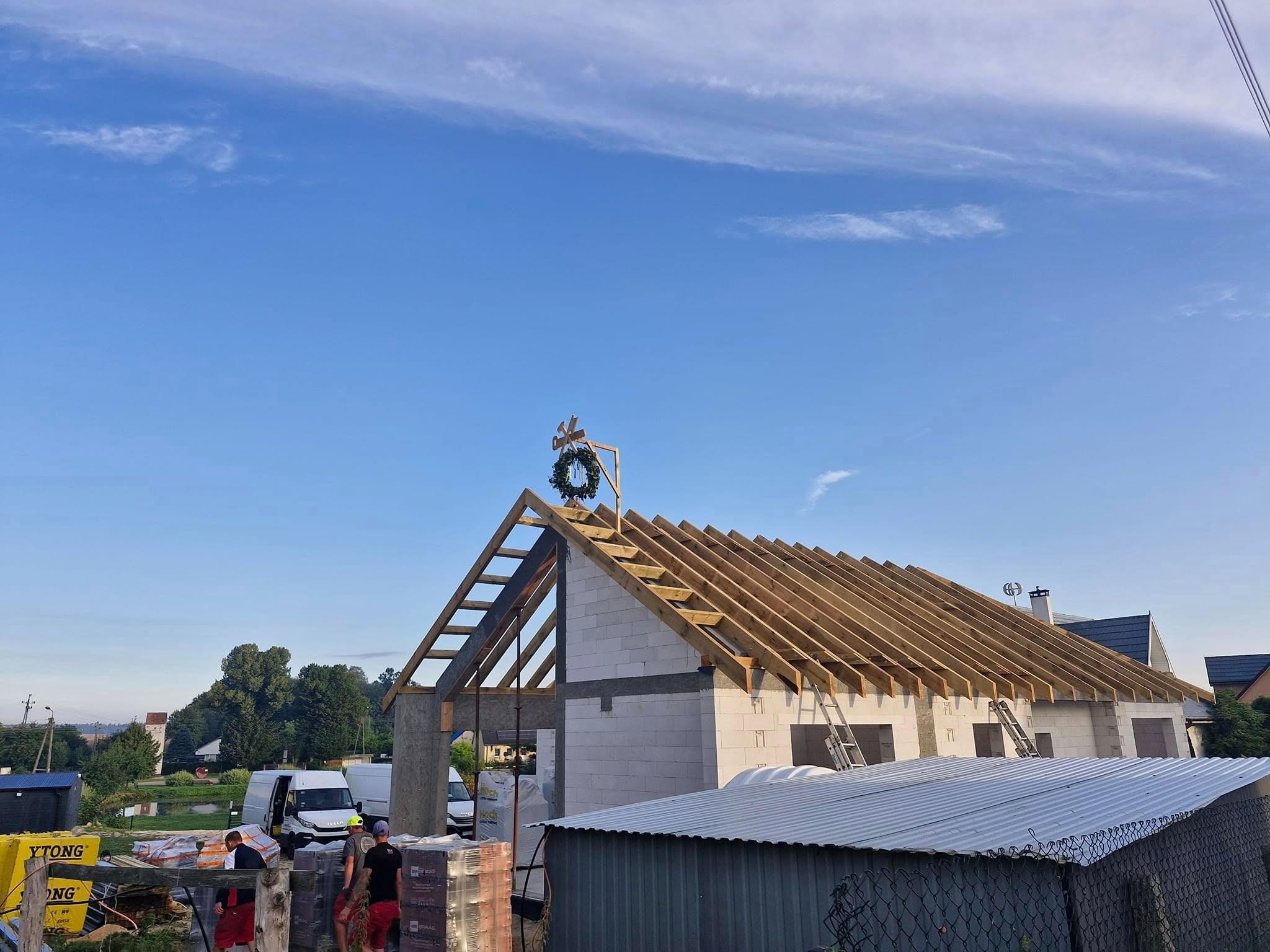 Wieniec na surowej więźbie dachowej domu z białego bloczka. W tle błękitne niebo i zabudowa. Częściowo widoczne materiały budowlane i pracownicy.
