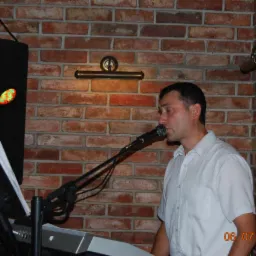 Mężczyzna śpiewa do mikrofonu, grając na keyboardzie Yamaha. W tle ceglana ściana i oświetlenie sceniczne. Data na zdjęciu: 06.07.2014.