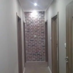 Wąski korytarz z ceglaną ścianą na końcu, oświetlony dwoma punktowymi lampami sufitowymi, z drewnopodobną podłogą i drzwiami po obu stronach.