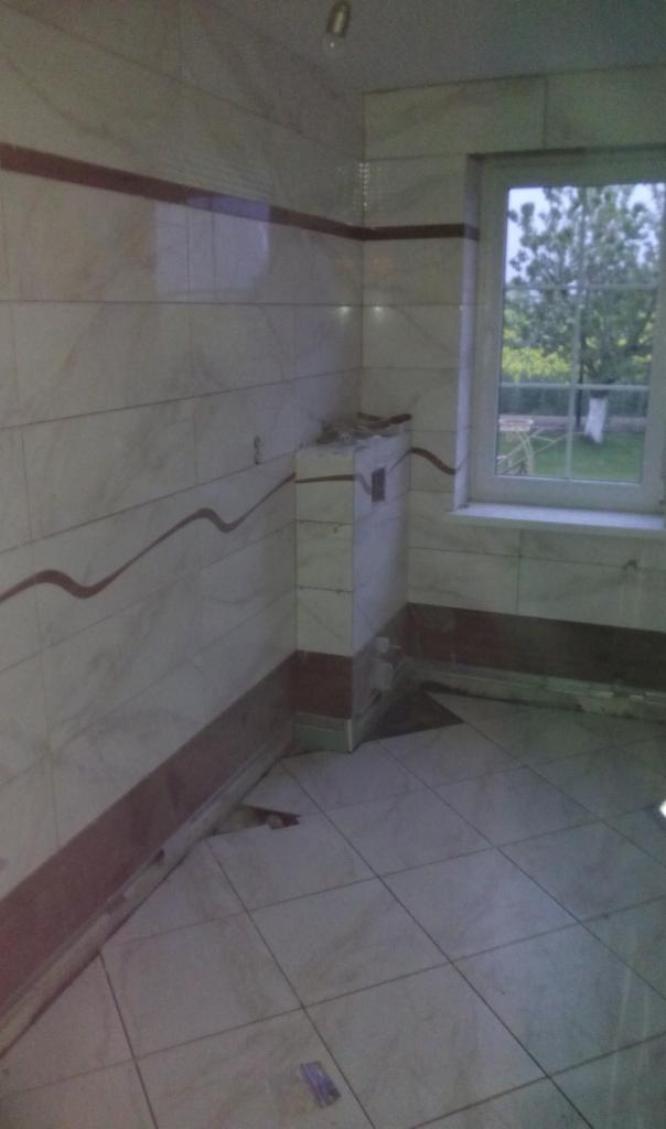 Wnętrze łazienki w trakcie prac wykończeniowych, widoczne białe płytki imitujące marmur z bordową dekoracyjną linią, częściowo obudowany element instalacji, okno z widokiem na zieleń.