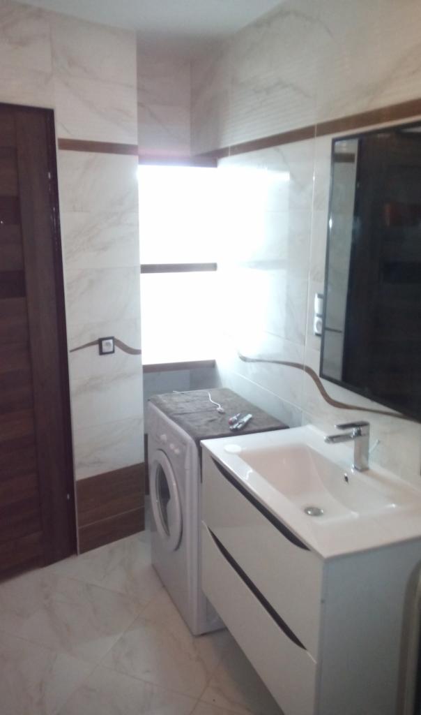 Wykończona łazienka z biało-beżowymi marmurkowymi płytkami, białą szafką z umywalką i pralką pod oknem.