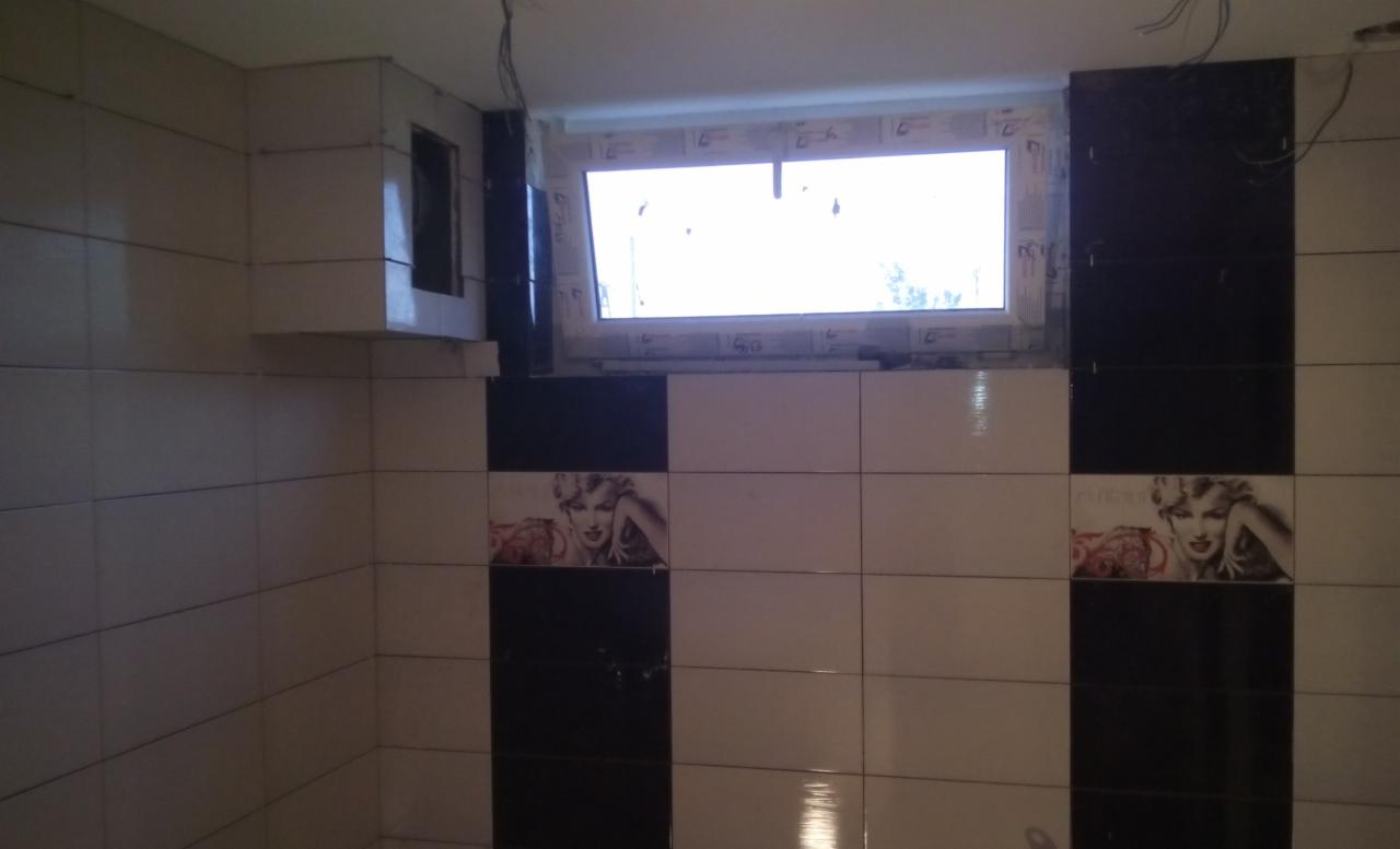 Ściana łazienki wyłożona biało-czarnymi płytkami z motywem Marilyn Monroe, widoczne okno z plastikową ramą i wystające kable ze ściany.