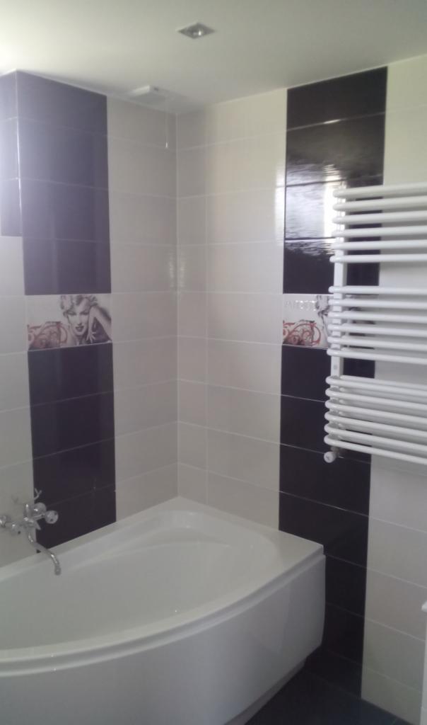 Biała narożna wanna z chromowaną baterią, ściany wyłożone biało-czarnymi płytkami z motywem Marilyn Monroe, grzejnik drabinkowy.