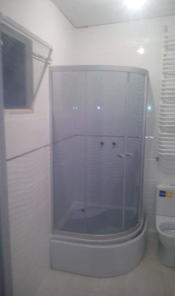 Nowa kabina prysznicowa z zaokrąglonym frontem i przesuwnymi drzwiami w jasnej łazience z białymi płytkami i grzejnikiem drabinkowym.