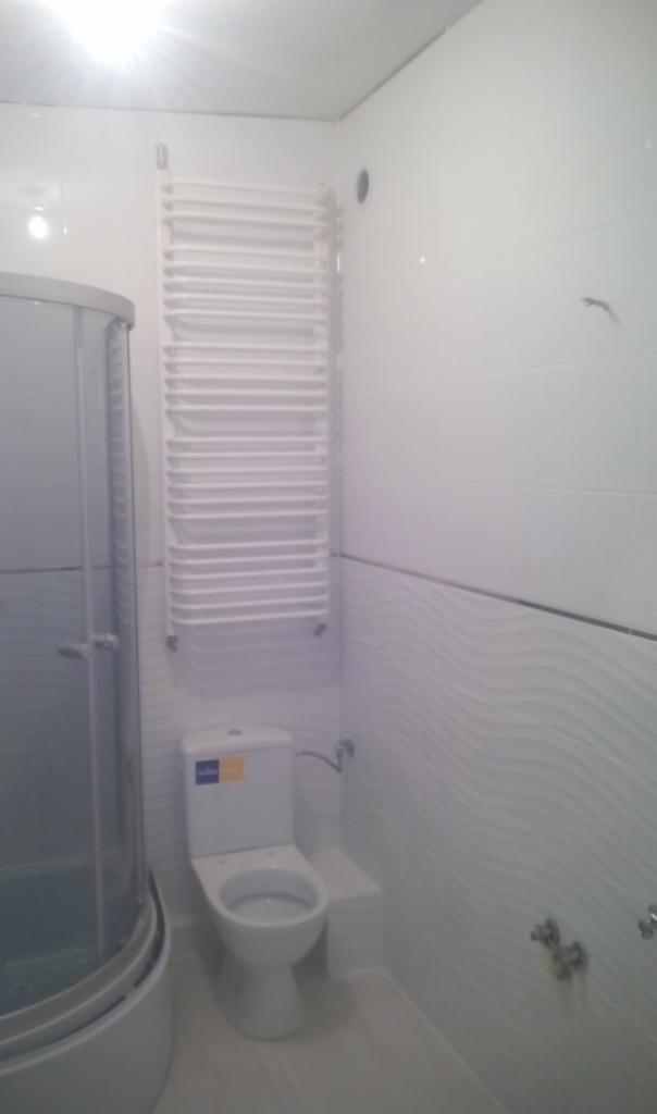 Biała łazienka z kabiną prysznicową, toaletą i grzejnikiem drabinkowym na ścianie z falistą fakturą płytek.