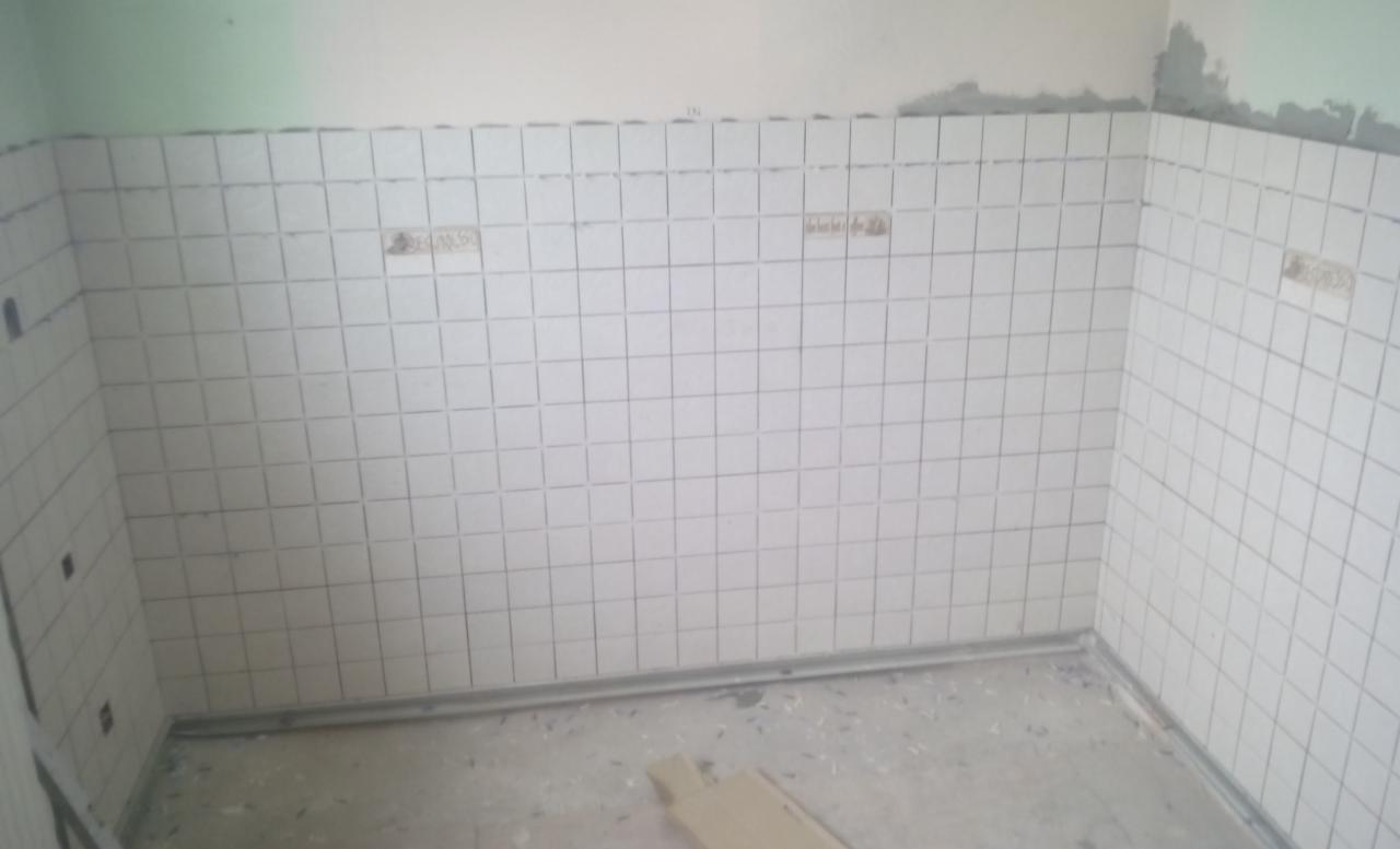 Ściany wyłożone białymi, kwadratowymi płytkami ceramicznymi, częściowo z widocznymi śladami kleju, w trakcie układania, z widocznym fragmentem podłogi i przygotowaną przestrzenią pod sufitem.