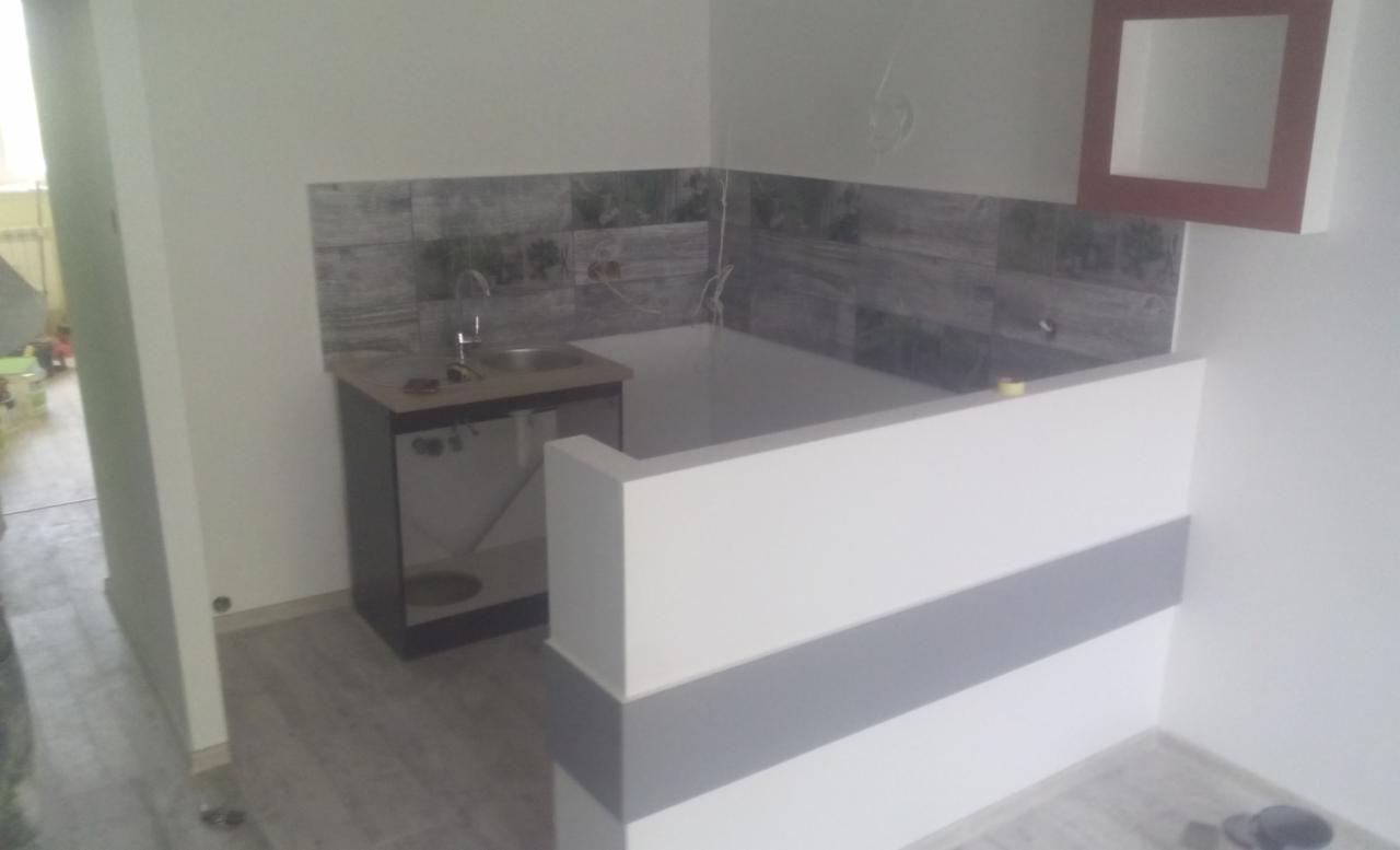 Fragment łazienki w trakcie wykańczania: umywalka z szafką, częściowo obłożona płytkami ściana imitująca drewno, niska ścianka działowa z białymi i szarymi pasami, oraz półka w ramce na ścianie.