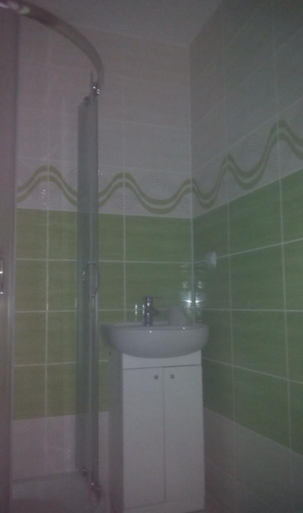 Umywalka z szafką w łazience wyłożonej zielono-białymi płytkami z dekoracyjnym paskiem, widoczny fragment kabiny prysznicowej z metalowym uchwytem.