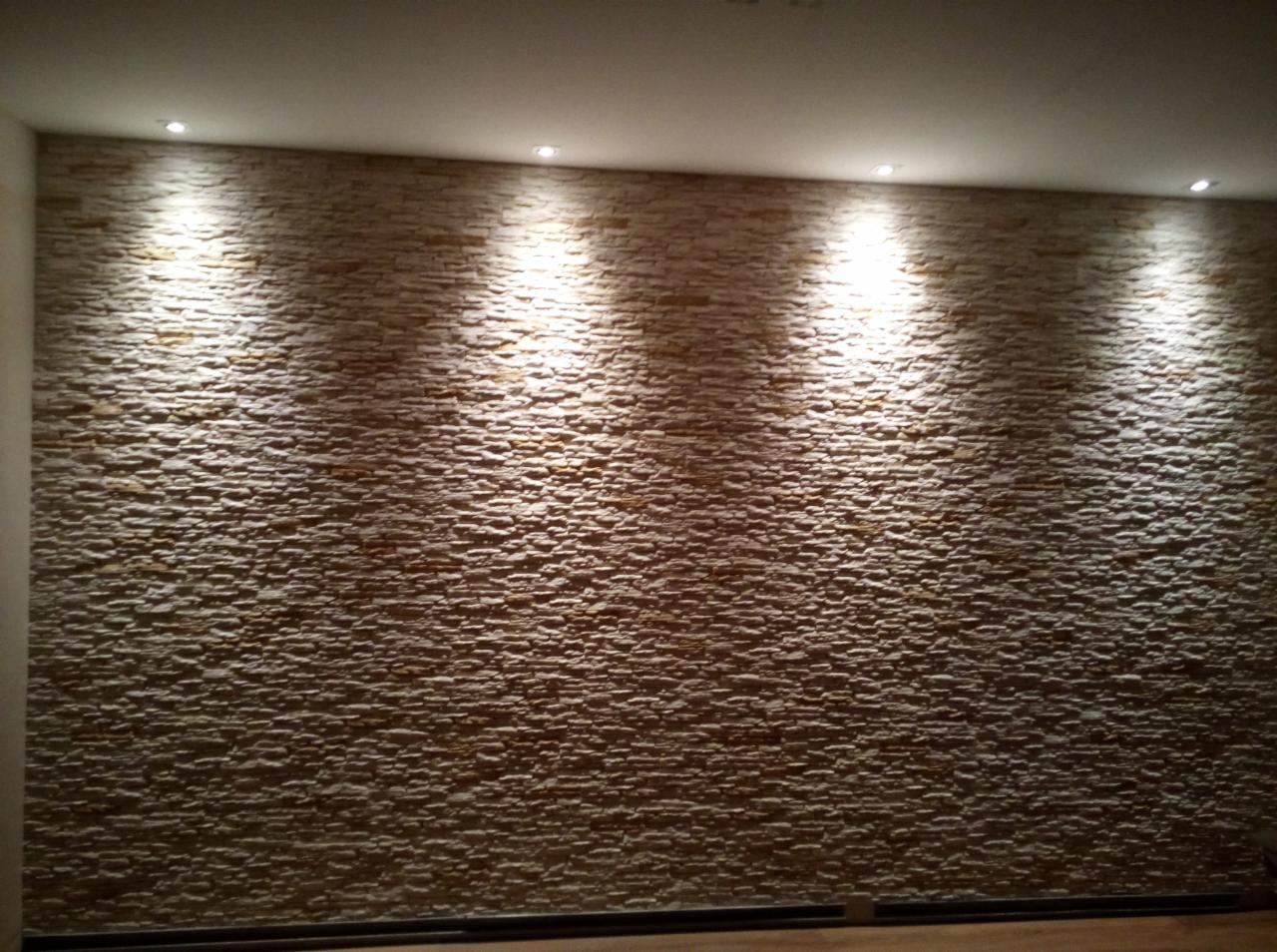Ściana wykończona dekoracyjnym kamieniem w odcieniach beżu i brązu, oświetlona punktowymi lampami sufitowymi, tworząca efekt trójwymiarowej tekstury.