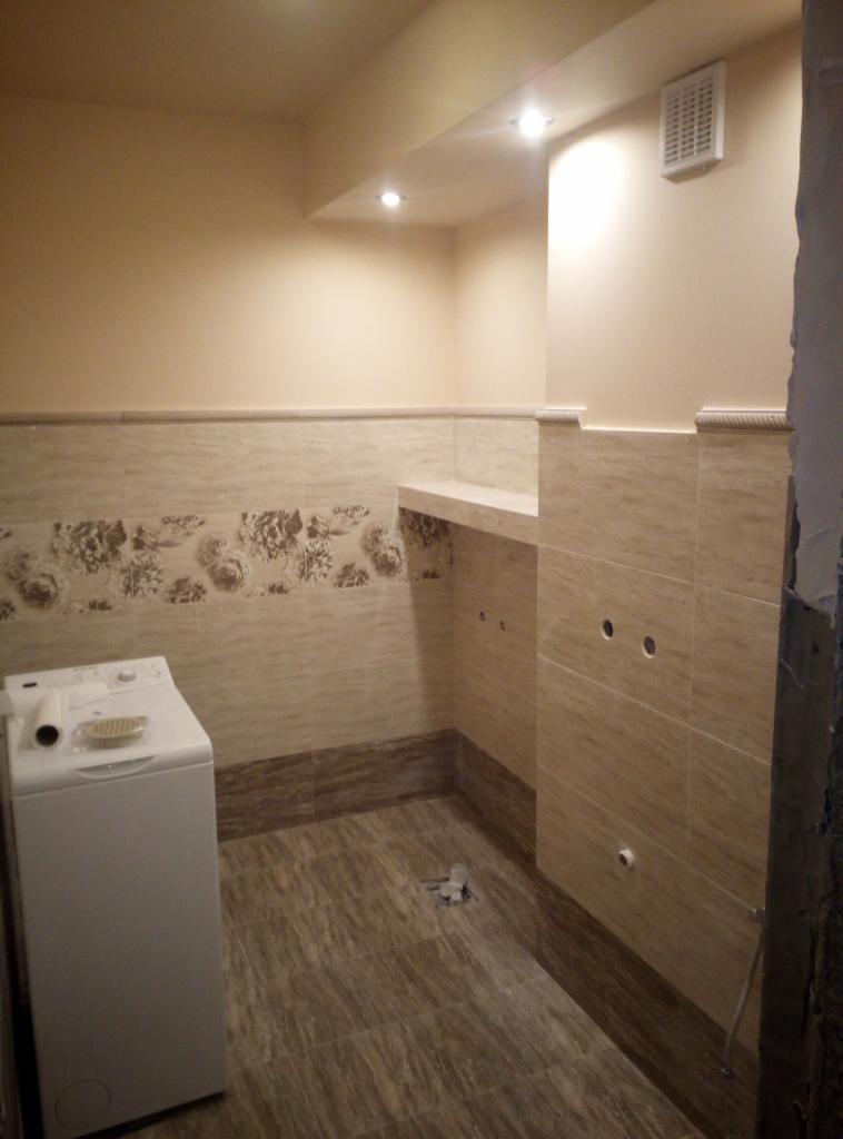 Łazienka po remoncie z pralką, beżowe płytki z kwiatowym motywem, widoczne otwory na instalacje, sufit z oświetleniem punktowym.