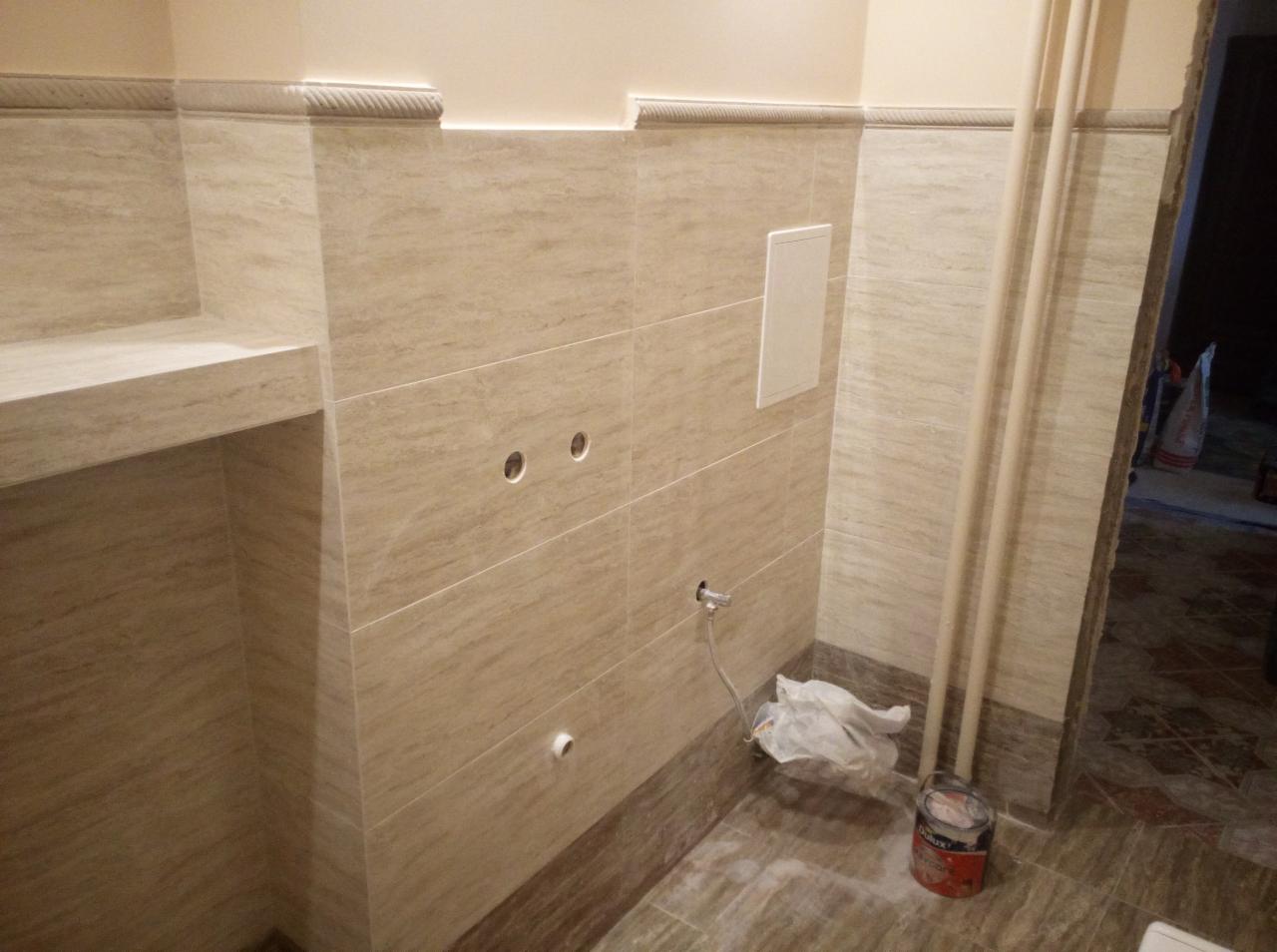 Łazienka w trakcie wykańczania: beżowe płytki imitujące marmur, otwory na armaturę, rury grzewcze przy ścianie, puszka farby Dulux na podłodze.