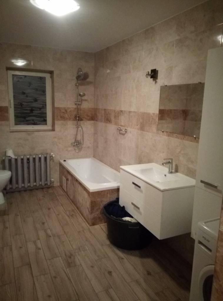 Ukończona łazienka z beżowymi płytkami, drewnopodobną podłogą, białą szafką z umywalką, wanną z prysznicem i grzejnikiem żeberkowym