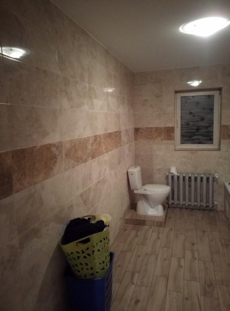 Wykończona łazienka z beżowymi płytkami, grzejnikiem żeberkowym i toaletą, widoczny kosz na pranie