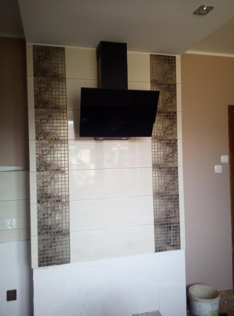 Czarny okap kuchenny zamontowany nad płytą wyłożoną beżowymi płytkami z dekoracyjnymi pasami mozaiki, widoczny fragment ściany pomalowanej na beżowo i wiadro na podłodze.