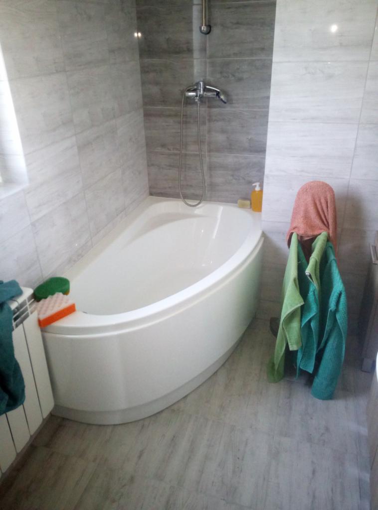 Biała wanna narożna z szarymi płytkami imitującymi drewno na ścianach i podłodze; widoczna bateria prysznicowa, grzejnik drabinkowy i ręczniki.