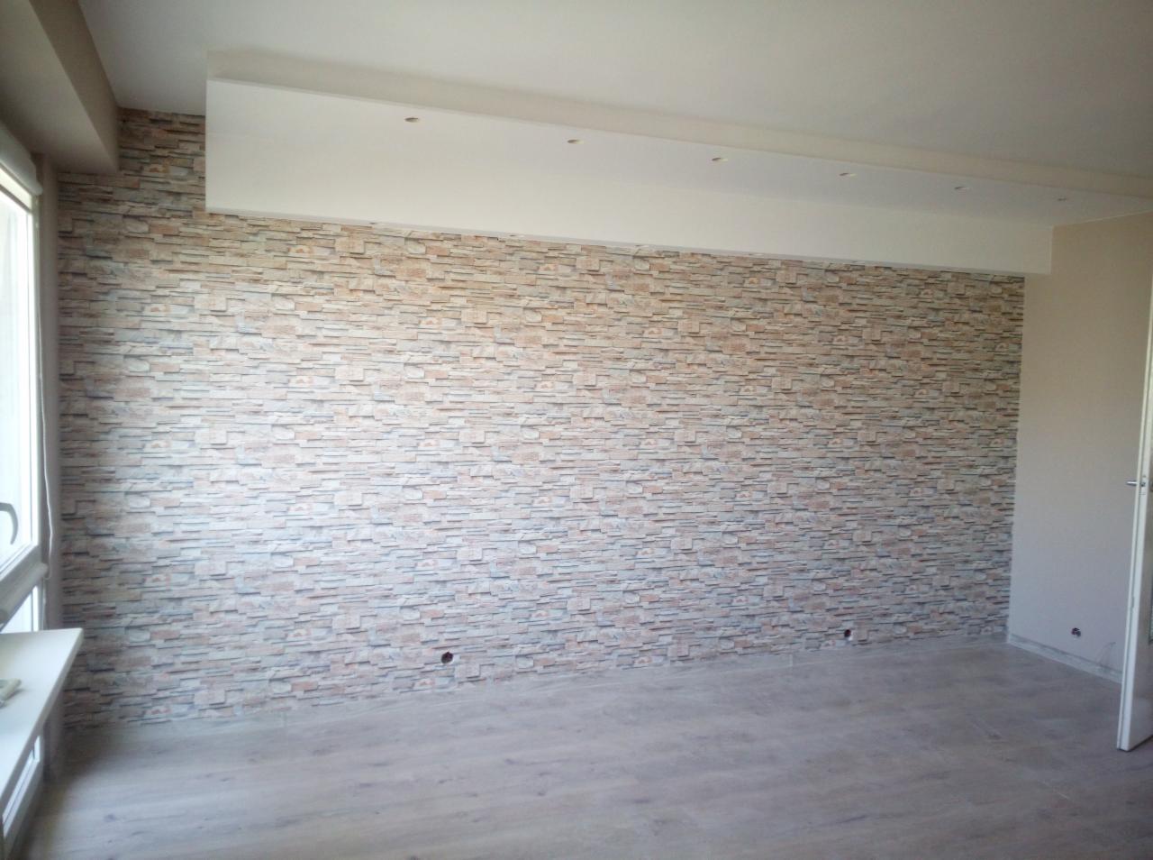 Ściana wykończona panelami imitującymi kamień w pustym pokoju z drewnianą podłogą i białym sufitem z wbudowanym oświetleniem.