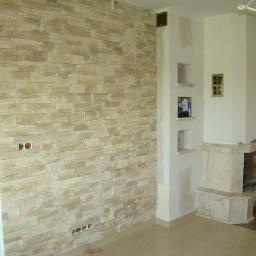 RAF-BUD Usługi Remontowo Budowlane - Wykończona ściana z dekoracyjnego kamienia w odcieniach beżu, z widocznymi gniazdkami elektrycznymi. W tle kominek z elementami dekoracyjnymi.