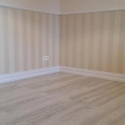 RAF-BUD Usługi Remontowo Budowlane - Jasne, pionowe pasy na ścianie w odcieniach beżu i bieli, wykończone białą listwą przypodłogową i drewnopodobną podłogą. Widoczne gniazdko elektryczne.