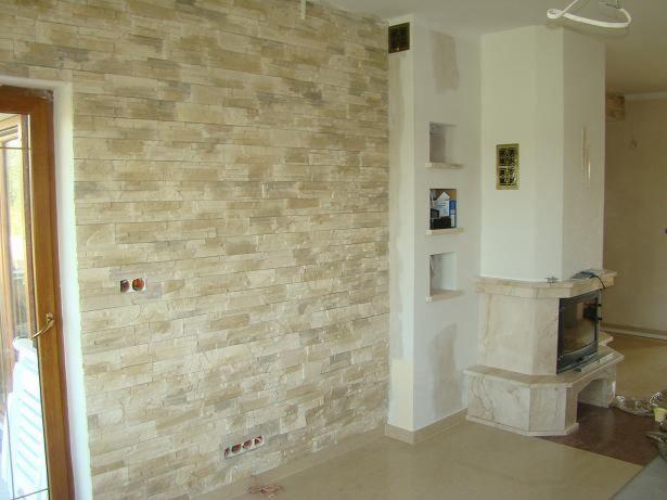 Wykończona ściana z dekoracyjnego kamienia w odcieniach beżu, z widocznymi gniazdkami elektrycznymi. W tle kominek z elementami dekoracyjnymi.