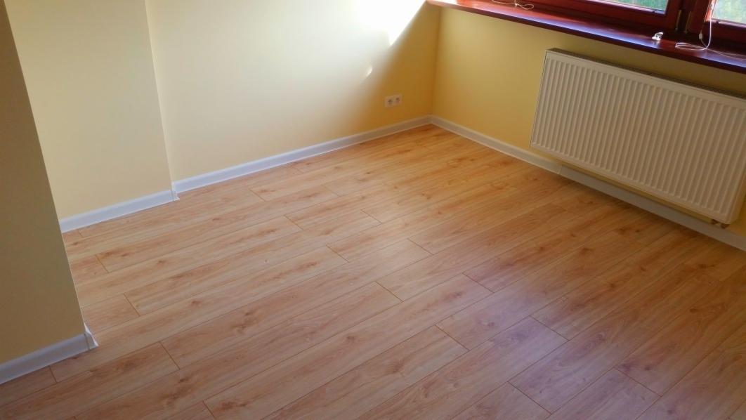 Jasny pokój po remoncie: panele podłogowe o wzorze drewna, żółte ściany z białymi listwami przypodłogowymi, grzejnik pod oknem. Schludne wykończenie.