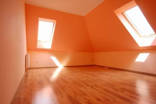 Pomarańczowe, puste poddasze z dwoma oknami dachowymi i parkietem. Świeżo pomalowane ściany i brak mebli sugerują gotowość do aranżacji.