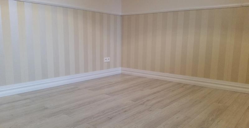 Jasne, pionowe pasy na ścianie w odcieniach beżu i bieli, wykończone białą listwą przypodłogową i drewnopodobną podłogą. Widoczne gniazdko elektryczne.