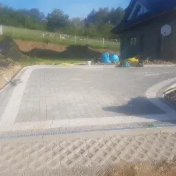 Ułożona szara kostka brukowa z białym obramowaniem na podjeździe, widoczne korytka odwadniające i ażurowe płyty w gruncie, w tle dom z anteną satelitarną i skarpa z zielenią.