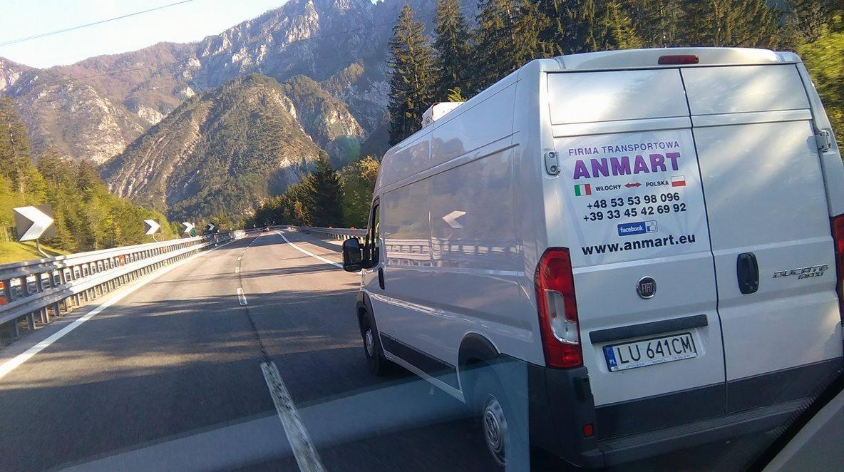 Bus transportowy firmy Anmart na tle górskiego krajobrazu. Bus w ruchu na drodze, widoczne numery kontaktowe i strona internetowa firmy na pojeździe.