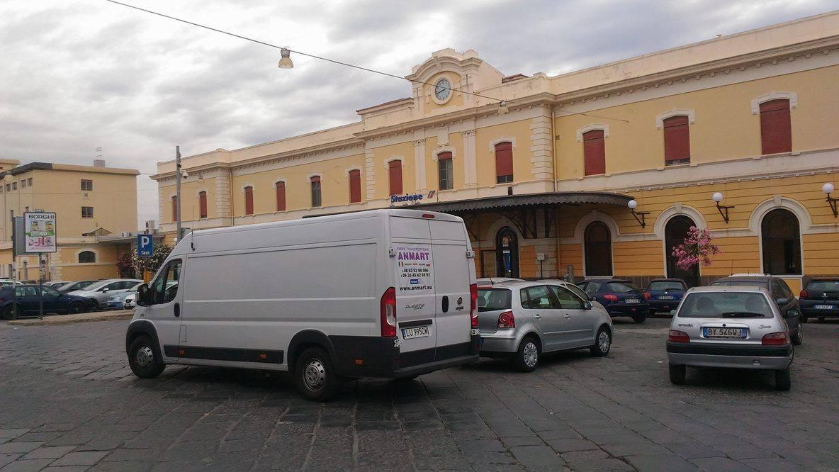 Biały bus firmy transportowej Anmart zaparkowany na tle budynku stacji kolejowej Stazione. Widoczne inne samochody i fragment parkingu.