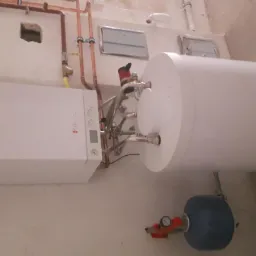 Zestaw grzewczy: biały piec gazowy Viessmann, zasobnik ciepłej wody i naczynie przeponowe na tle surowej ściany. Miedziane rury instalacji.