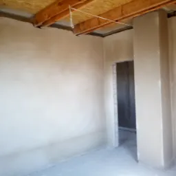 Wnętrze w stanie surowym po nałożeniu jasnej warstwy tynku na ściany, widoczne drewniane elementy konstrukcyjne pod sufitem i fragment otworu drzwiowego