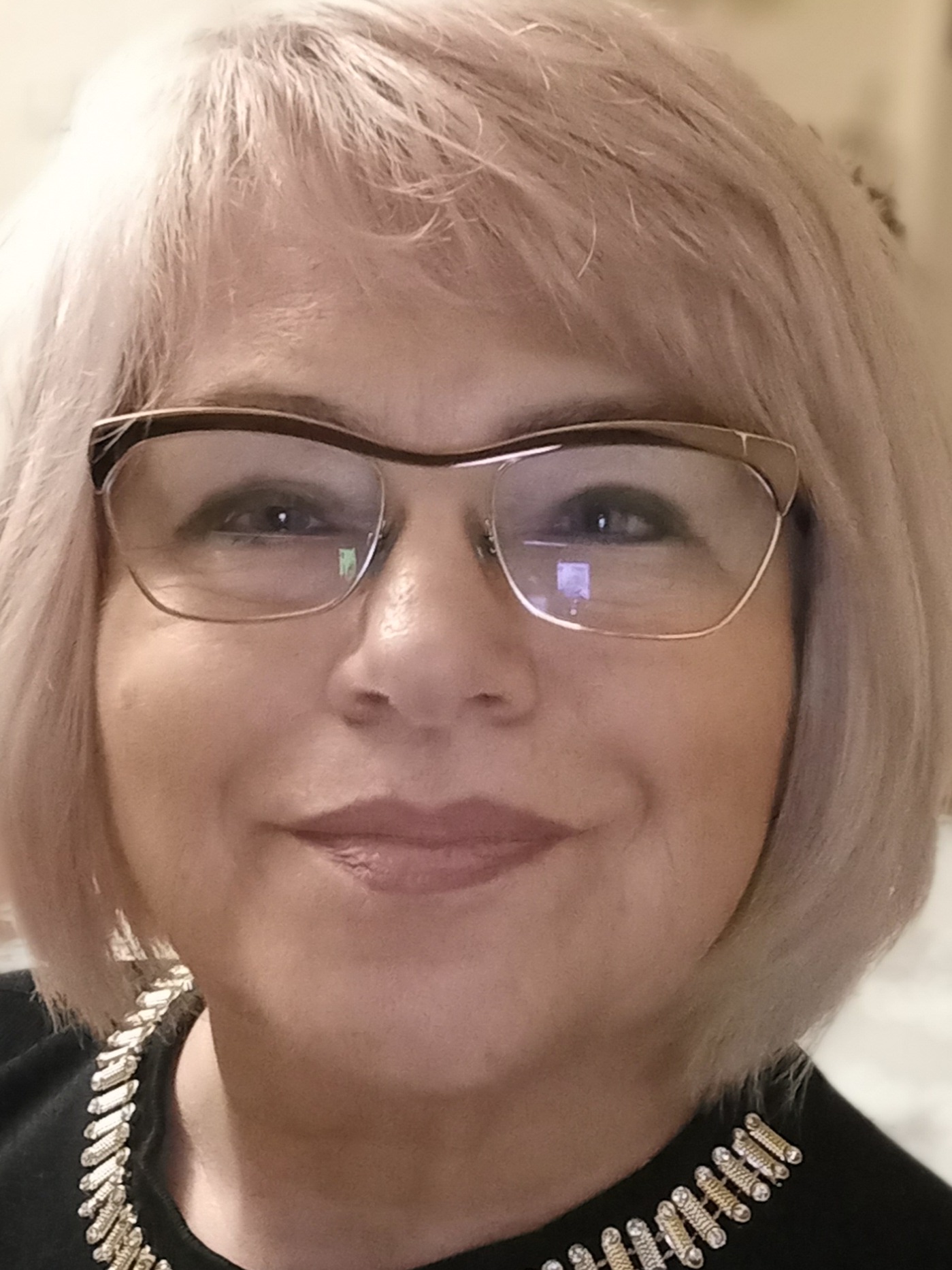 Portret kobiety w okularach i czarnej bluzce z ozdobnym kołnierzem. Krótkie, jasne włosy. Uśmiech. Widoczne odbicie światła w szkłach okularów.