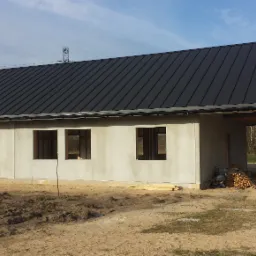 Surowy, parterowy budynek w trakcie budowy z ciemnym, metalowym dachem, widoczne otwory okienne i drzwiowe, na zewnątrz stos drewna, w tle drzewa i słup energetyczny.