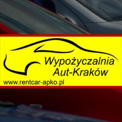 Żółty baner z czarnym napisem 'Wypożyczalnia Aut-Kraków' i adresem strony na tle zaparkowanych aut w odcieniach czerwieni i granatu, częściowo poza kadrem.