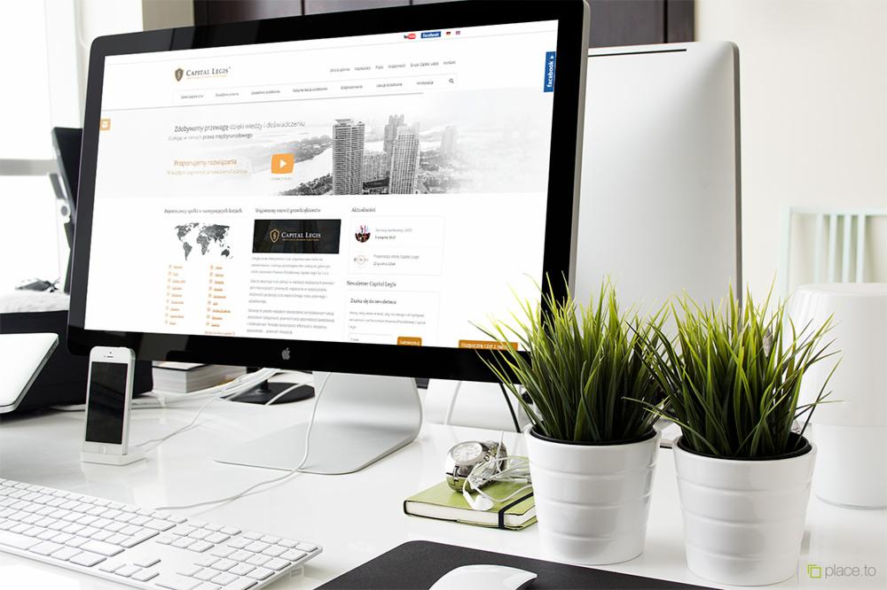 Monitor wyświetlający stronę internetową firmy Capital Legis, widoczny na biurku w Deszcznie obok telefonu, klawiatury, okularów i dwóch doniczek z trawą.